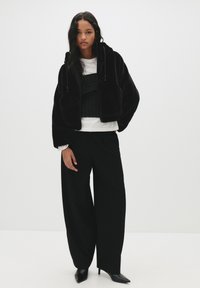 Črna umetna krzna jakna s kapuco, z zadrgo in vrvicami, v kombinaciji s črnim oprijetim topom in širokimi črnimi hlačami, dopolnjena s špicastimi petami.