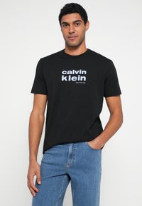 Czarny bawełniany t-shirt z białym napisem „calvin klein” oraz „New York City”, okrągły dekolt, krótki rękaw i luźny krój. Widoczne niebieskie jeansy.