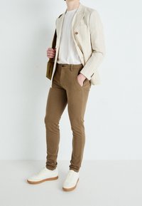 Béžová pruhovaná sako, bílé tričko, hnědé slim-fit kalhoty a bílé tenisky s gumovými podrážkami. Minimalistický design s jemnými detaily.
