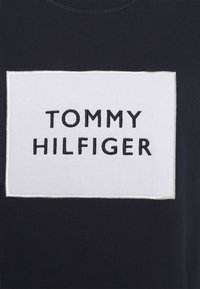 Tessuto nero con una toppa rettangolare bianca ricamata con "TOMMY HILFIGER" in lettere maiuscole nere.