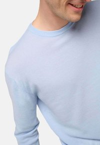 Mann lächelt und trägt einen hellblauen Rundhals-Pullover aus strukturiertem Strickstoff vor einem weißen Hintergrund.