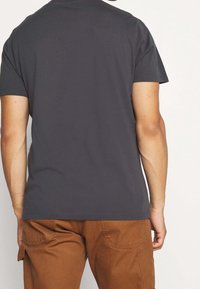 Ciemnoszary bawełniany t-shirt z krótkimi rękawami i okrągłym dekoltem, o gładkiej fakturze. Noszony z brązowymi spodniami bojówkami, z widocznymi kieszeniami.