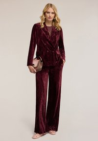Bordowy aksamitny garnitur z podwójnym zapięciem, szerokimi spodniami i różowymi balerinami oraz małą torebką.