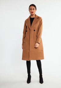 Cappotto lungo marrone doppiopetto con rever a falde, tasche frontali e cuciture strutturate, indossato sopra leggings neri e stivali con tacco alto.