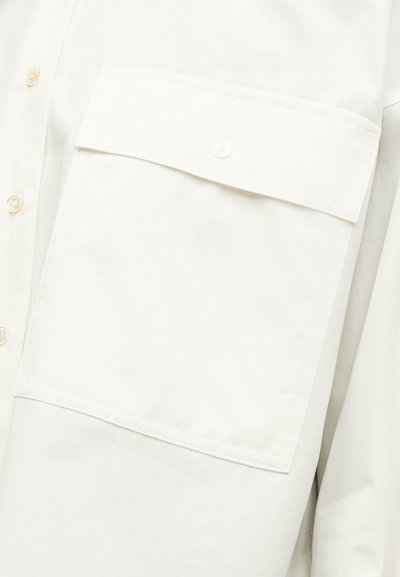 Κοντινό πλάνο μπροστινού μέρους λευκού πουκαμίσου με μεγάλη τσέπη στο στήθος με κουμπιά και ορατή ραφή σε λείο ύφασμα.