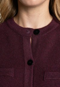 Burgunderfarbener Strickcardigan mit gerippter Textur, versehen mit drei schwarzen Knöpfen, einem runden Halsausschnitt und zwei kleinen aufgenähten Taschen.