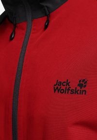 Veste rouge à texture lisse, dotée d'une fermeture éclair noire et d'une capuche, avec un logo noir sur le côté gauche de la poitrine. Conçue pour les vêtements d'extérieur.