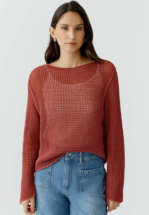 Femme portant un pull en maille ample à manches longues de couleur rouille sur un débardeur et un jean bleu taille haute, sur un fond uni.