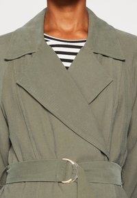 Oliwkowy płaszcz trencz z szerokim kołnierzem, paskiem w talii z złotym pierścieniem oraz delikatnymi detalami przeszyć. Pod spodem w paski top.