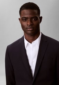 Νέος μαύρος άνδρας που φοράει μαύρο σακάκι και λευκό πουκάμισο με γιακά, κοιτάζει μπροστά με ουδέτερη έκφραση σε γκρι φόντο.