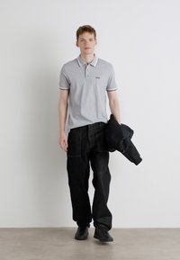 Ανοιχτό γκρι πόλο με γιακά, μαύρες λεπτομέρειες; συνδυάζεται με χαλαρές μαύρες τζιν παντελόνες. Το μοντέλο κρατάει μια μαύρη ζακέτα. Μαύρα παπούτσια.