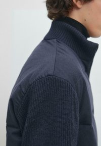 Boční profil osoby v námořnicky modré bundě s pletenými texturovanými rukávy a vroubkovaným vysokým límcem.