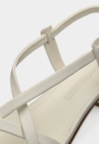 Κοντινή λήψη λευκού σανδαλιού με λουράκια, χαμηλό τακούνι και λείο δερμάτινο φινίρισμα σε απλό φόντο.