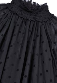 Schwarzes Tüllkleid mit Polka-Dots, das über ein gerafftes Oberteil und einen Volant-Ausschnitt verfügt. Der Stoff ist leicht und durchscheinend, was die Textur der Punkte betont.