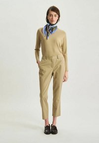 Haut beige à manches longues, pantalon beige ajusté, écharpe en soie à motif bleu en complément, et mocassins en cuir noir.