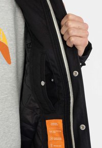 Giubbotto esterno nero con zip e chiusure a scatto, dotato di un interno trapuntato, tasche laterali e un'etichetta per prestazioni arancione all'interno.