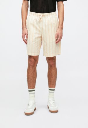 Lys ferskenfargede shorts med vertikale kremstriper, med elastisk midje og snøring. Kombinert med hvite sneakers og stripete sokker.