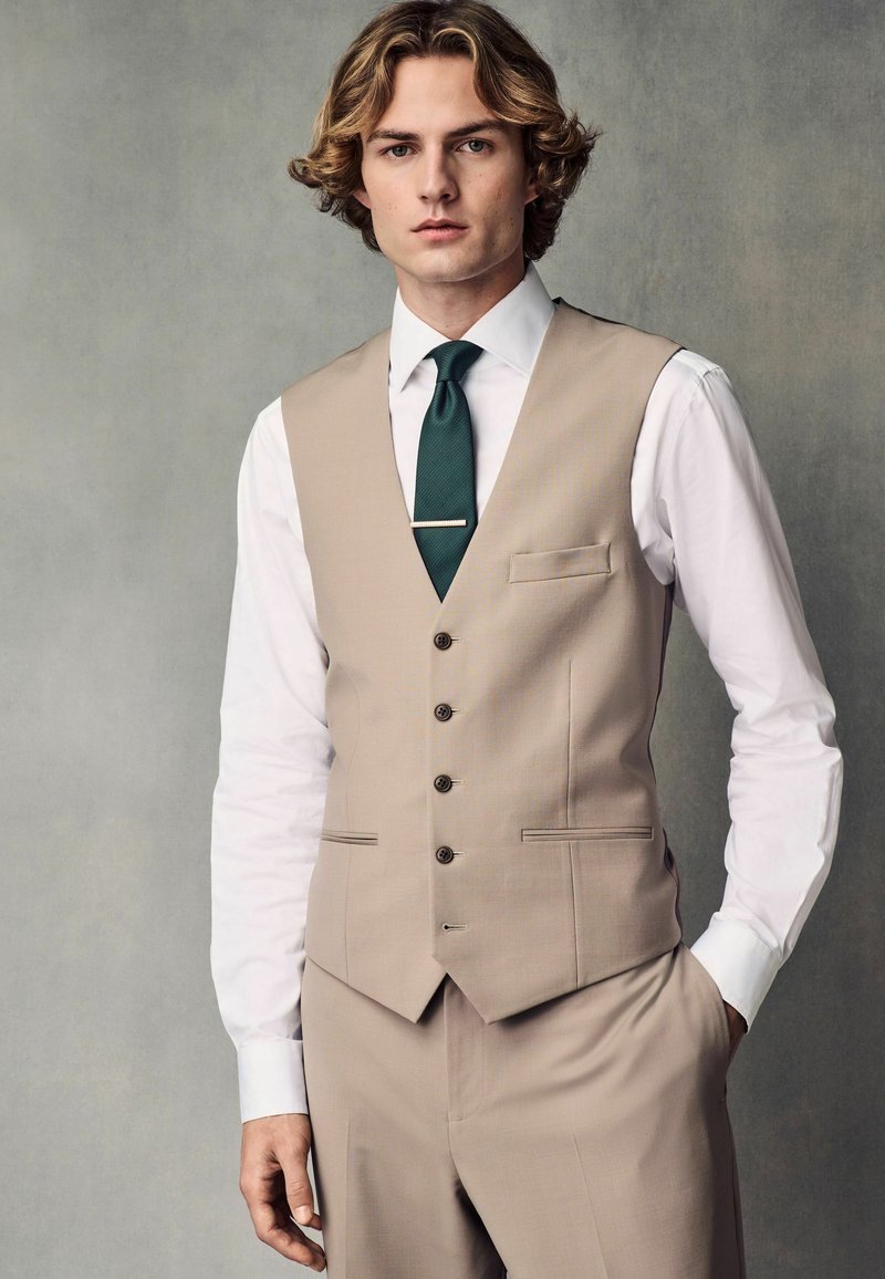 Gilet beige avec un col en V et cinq boutons, associé à une chemise blanche et une cravate vert foncé. Coupe ajustée, texture lisse et détails de poche latérale.