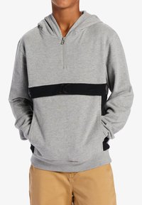 Grå huvtröja med halvpull, svart elastisk band vid midjan, kängurufickor och ribbade mudd, kombinerat med khakifärgade byxor.