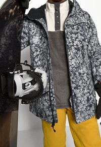 Snowboardová bunda v černobílém květinovém vzoru, šedá fleecová středn�í vrstva, žluté kalhoty, držící snowboard a vázání.