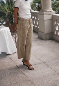 Jeans anchos color caqui con pliegues frontales y bolsillos, combinados con una blusa blanca que tiene un cuello negro, y sandalias de tacón alto y tiras negras.