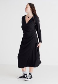 Robe noire à manches longues, design drapé et texture douce, associée à des baskets montantes noires et des chaussettes mi-mollet grises.