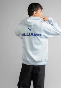 Mężczyzna w jasnoniebieskiej bluzie Puma z napisem "WILLIAMS" na plecach, w połączeniu z czarnymi spodniami, na jednolitym szarym tle.