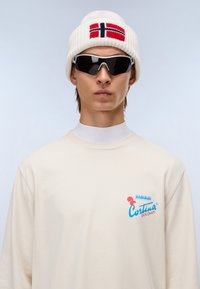 Μπεζ πουκάμισο με μακριά μανίκια, φέρει μπλε και κόκκινο λογότυπο "Cortina Dolomiti", σε συνδυασμό με λευκό πλεκτό σκουφάκι που διαθέτει κόκκινη και μπλε νορβηγική σημαία, καθώς και σκούρα γυαλιά ηλίου.