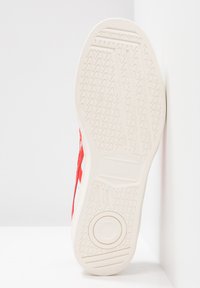 Röd och vit sneaker med texturerad gummisula, som har ett vågigt mönster och en cirkulär emblem på undersidan. Sidan har en kontrasterande design.