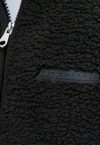 Chaqueta de forro polar negra con superficie texturizada, que incluye un cierre frontal con cremallera y un parche con el logotipo negro elevado en el lado izquierdo.