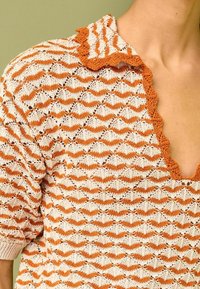 Gebreide top met een oranje en witte zigzagpatroon, voorzien van een gegolfde kraag en korte mouwen, gemaakt van een lichte, gestructureerde stof.