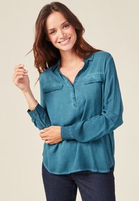 Blusa lunga a maniche larghe di colore turchese, realizzata in tessuto liscio, con una piastra a mezza bottone, due tasche al petto e un orlo curvo.