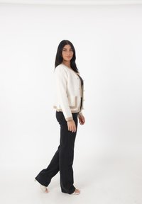 Cardigan bianco sporco con dettagli beige, chiusura a bottoni e tasche, abbinato a jeans neri a zampa d'elefante e sandali aperti. Sfondo bianco uniforme.