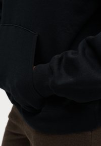 Main d'une personne dans la poche avant d'un sweat à capuche noir, montrant le poignet de la manche et une partie d'un pantalon marron.