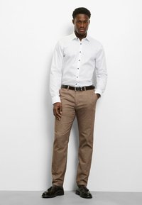 Weißes Langarmhemd mit schwarzen Knöpfen, kombiniert mit hellbraunen, strukturierten Hosen und schwarzen Schuhen mit glänzendem Finish.