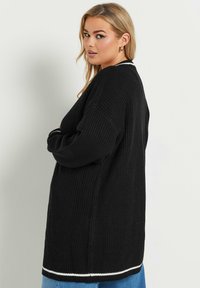 Svart stickad cardigan med ribbat mönster, med en vit accent längs fållen och en lös, oversized design.