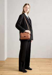 Sac en cuir tan à bandoulière avec un design structuré, des accessoires en or et un rabat à l'avant. Porté avec un blazer noir à rayures et un pantalon noir.