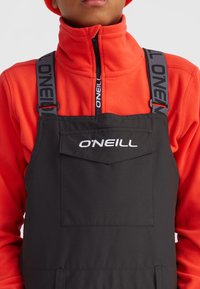 Čierne overall s nastaviteľnými popruhmi, na ktorých sa nachádza logo "O'NEILL", nosené cez jasne oranžový fleece vrch. Obsahuje detail prednej vrecka.