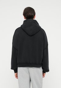 Hoodie preto de tamanho oversized com capuz ajustável, ombros caídos e detalhes de bainha desfiada, usado sobre calças de moletom cinzentas claras.
