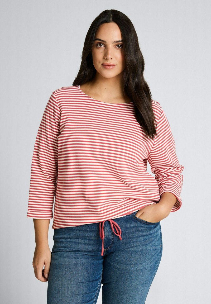 Junge Frau mit langen dunklen Haaren, die ein langärmliges, rot-weiß gestreiftes Shirt und eine blaue Jeans trägt, eine Hand in der Tasche, blickt nach vorne.