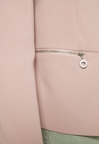 Veste rose pâle en tissu texturé, dotée d'une fermeture éclair argentée, d'un anneau circulaire en accent et d'une finition lisse.