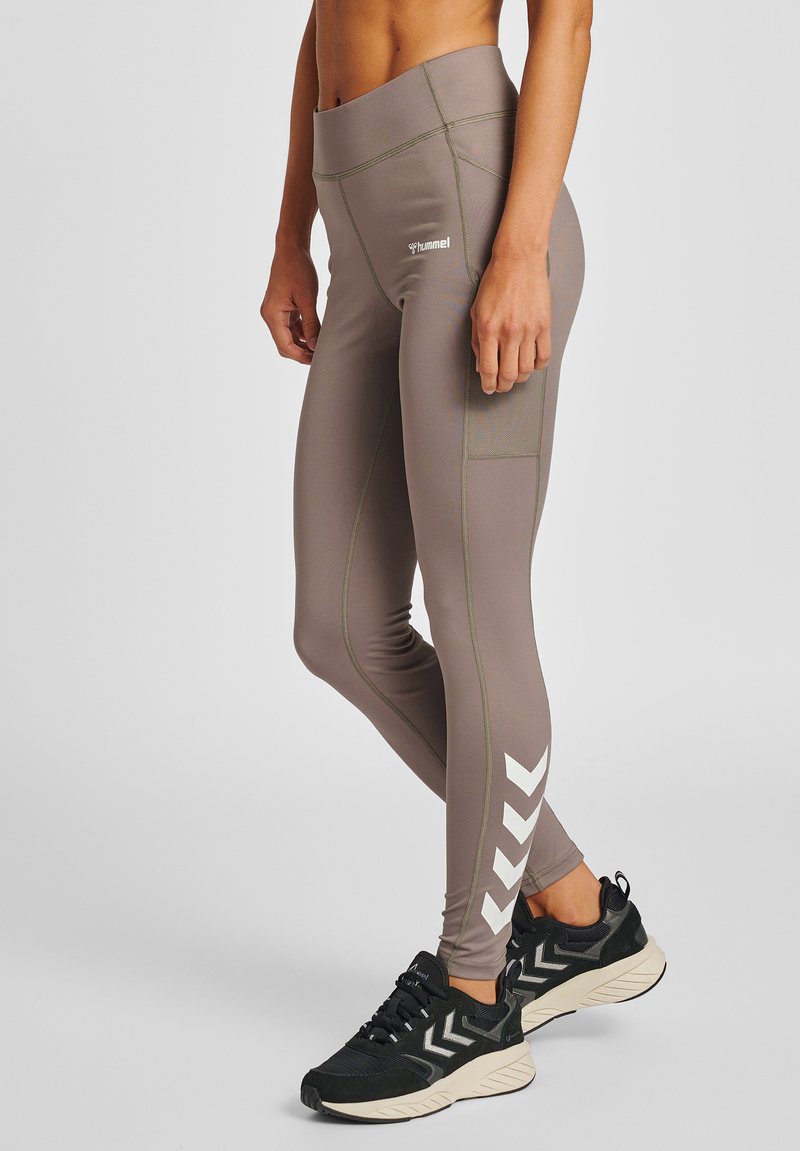 Sportleggings in Taupe mit weißen Chevron-Details an den Waden, Netz-Tasche am Oberschenkel, getragen mit schwarzen und beigen Sportschuhen.