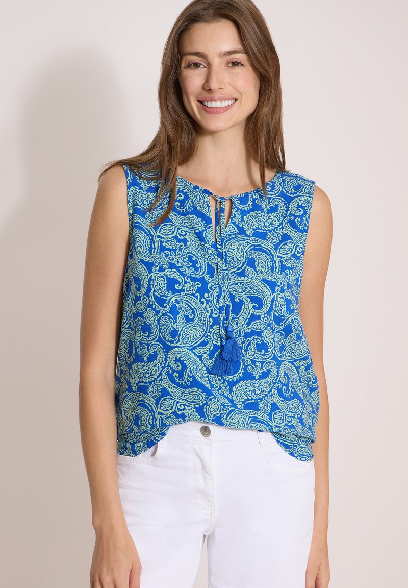 Femme souriante portant un haut bleu sans manches à motif paisley avec des liens à pompons et un pantalon blanc sur un fond clair uni.