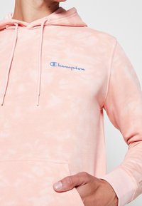 Sudadera con capucha de color rosa claro hecha de tela suave con un patrón tie-dye. Cuenta con un bolsillo delantero, capucha con cordón y el logo "Champion" en azul.