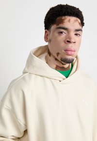 Svijetlosmeđa hoodie s opuštenim krojem, koja ima prednji kengur džep i mali bijeli logo na lijevoj strani prsnog koša. Mekana tekstura tkanine.