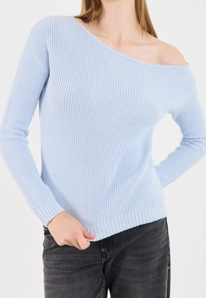 Femme portant un pull tricoté bleu clair épaules dénudées à manches longues et un jean noir, main tenant l'ourlet du pull.