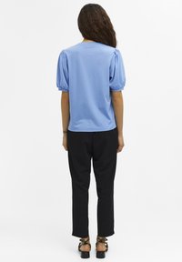 Frau mit langen lockigen Haaren, die ein blaues Top mit Puffärmeln, schwarze Hosen und schwarze Sandalen trägt, stehend mit dem Rücken zu einer weißen Hintergrund.