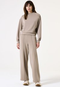 Beige Sweatshirt mit hohem Kragen und seitlichem Reißverschluss, kombiniert mit lockeren, hochgeschnittenen beige Hosen. Glatter Stoff, minimalistisches Design.