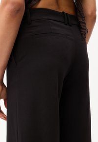 Pantalon noir à coupe ajustée, présentant une texture lisse, une taille fine et un détail de poche arrière décoratif.