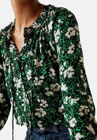 Kvinde iført en langærmet bluse i grøn, sort og hvid med blomsterprint, knaplukning foran og flæsekrave, parret med blå jeans.